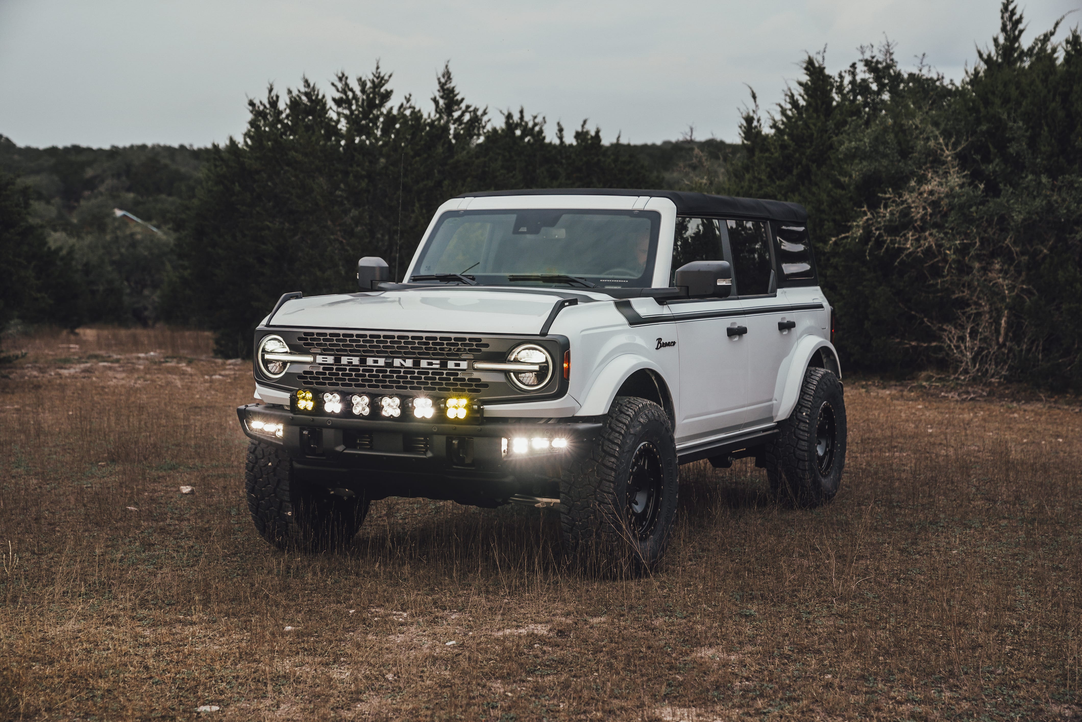 2021 Ford Bronco Badlands in Oxford White with Baja Design Fog Light Pocket Kit, XL Squadron Sport Linkable Lights, 17" Raceline wheels, 35x12.50R17 Yokohama Geolander X-AT Tires, Rough Country Spare Tire Extender, Third Brake Light Relocation Bracket, Paint Matched Fender Flares, Retro Striping, Retro Badges